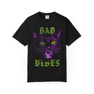 A flat lay image of a black t-shirt featuring a graphic of a purple cat's head with green eyes and green text reading BAD VIBES.