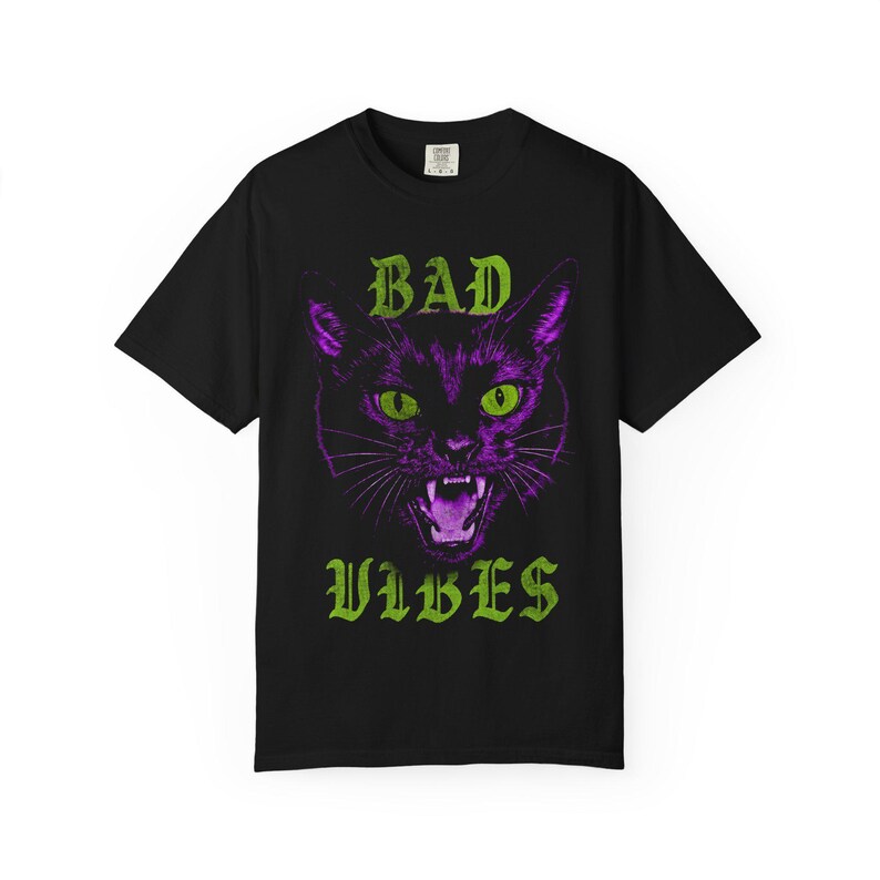 A flat lay image of a black t-shirt featuring a graphic of a purple cat's head with green eyes and green text reading BAD VIBES.