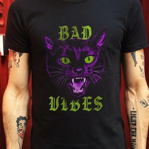 May include: A black short-sleeve t-shirt featuring a graphic of a snarling cat with purple fur and bright green eyes. The text "BAD" is printed in green above the cat, and the text "VIBES" is printed in green below the cat in a gothic font.