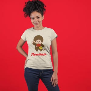 A woman with curly hair wearing a cream-colored t-shirt with a Monchhichi graphic and blue jeans stands against a red backdrop.