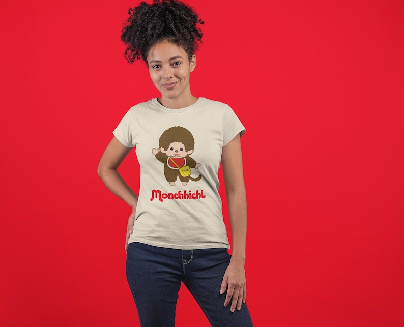 A woman with curly hair wearing a cream-colored t-shirt with a Monchhichi graphic and blue jeans stands against a red backdrop.