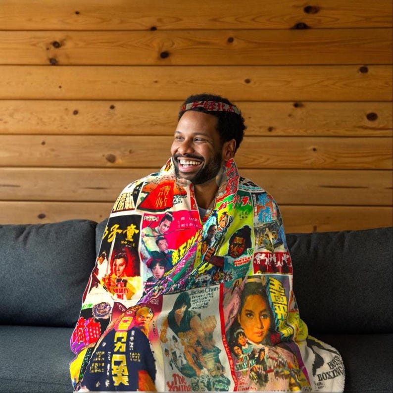 A man with a beard and headband sits on a gray sofa, smiling and wrapped in a blanket patterned with vintage Kung Fu movie posters.