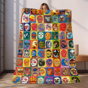 Op de afbeelding: Een kleurrijke deken met een raster van luchador-masker illustraties. De maskers zijn in verschillende kleuren en ontwerpen, tegen een mix van achtergronden. De deken wordt omhoog gehouden, wat het levendige kunstwerk laat zien.