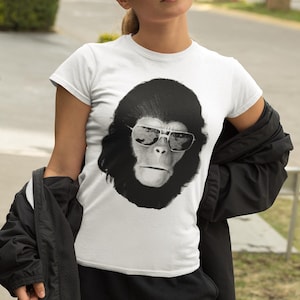 May include: White t-shirt with a black and white image of a chimpanzee wearing sunglasses. The graphic is centered on the shirt. The shirt has short sleeves and a crew neck. The person is wearing black trousers and a black jacket.