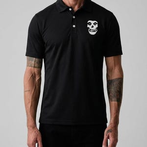A man wearing a black short-sleeved polo shirt with a white embroidered skull graphic on the left chest.