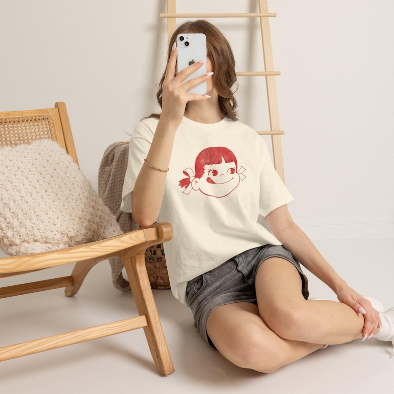 A person sits on a wooden floor, holding a phone to their face, wearing a cream-colored t-shirt with a red graphic of a girl's face with pigtails and a winking expression, paired with dark shorts.