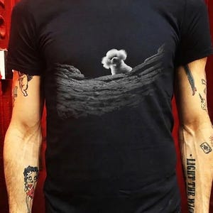 May include: A black short-sleeved t-shirt featuring a graphic print of a small white fluffy dog peeking over a textured, dark grey rock formation. The design is centred on the chest area of the garment, providing a clear contrast between the dog and the dark background.