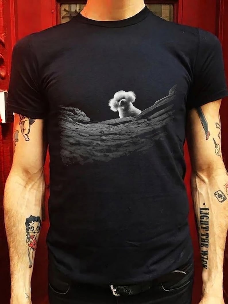 May include: A black short-sleeved t-shirt featuring a graphic print of a small white fluffy dog peeking over a textured, dark grey rock formation. The design is centred on the chest area of the garment, providing a clear contrast between the dog and the dark background.