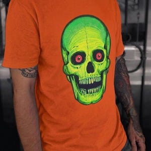 May include: A bright orange short-sleeved t-shirt featuring a large, centred graphic of a neon green skull. The skull has deep-set, dark red circular eyes and a wide, open mouth with visible teeth, rendered in a bold, illustrative style.