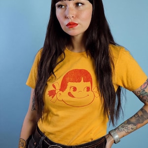 A person with long dark hair and red lipstick wears a mustard yellow t-shirt with a red graphic of a girl's face with pigtails and a winking expression, paired with black jeans and a belt, against a blue background.