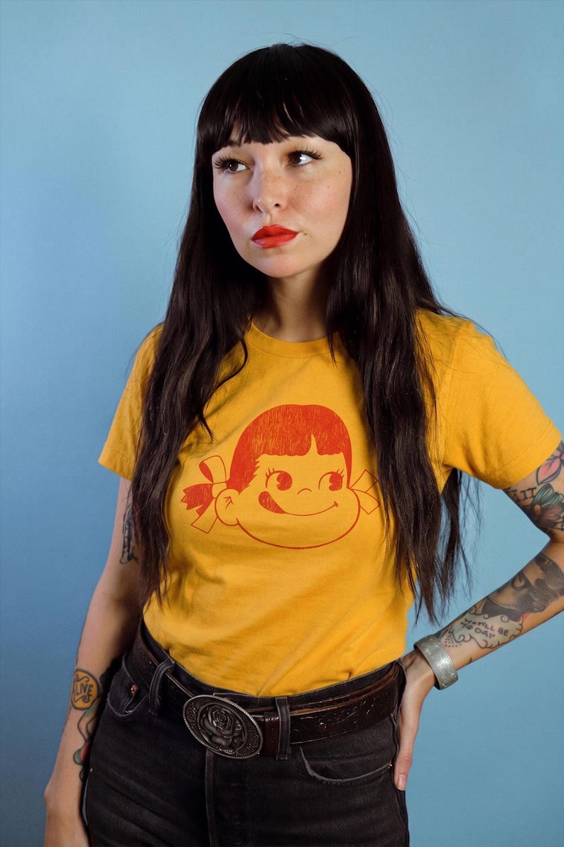 A person with long dark hair and red lipstick wears a mustard yellow t-shirt with a red graphic of a girl's face with pigtails and a winking expression, paired with black jeans and a belt, against a blue background.