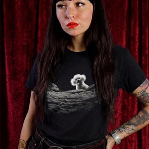 May include: A black short-sleeved t-shirt featuring a graphic print of a small white fluffy dog peeking over a textured, cratered lunar surface. The design is centred on the chest, contrasting the white dog against the dark grey moon landscape.