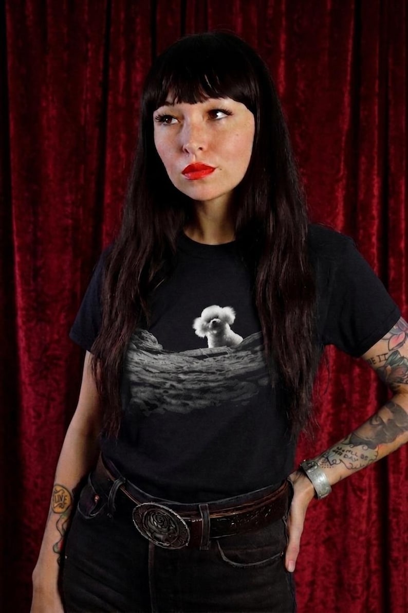 May include: A black short-sleeved t-shirt featuring a graphic print of a small white fluffy dog peeking over a textured, cratered lunar surface. The design is centred on the chest, contrasting the white dog against the dark grey moon landscape.