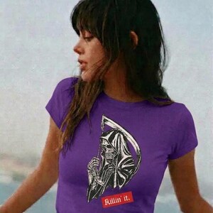 A person outdoors wearing a purple t-shirt featuring a black and white grim reaper graphic and red text.