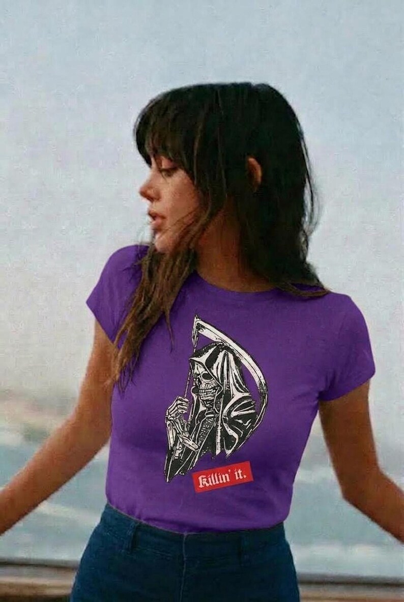 A person outdoors wearing a purple t-shirt featuring a black and white grim reaper graphic and red text.
