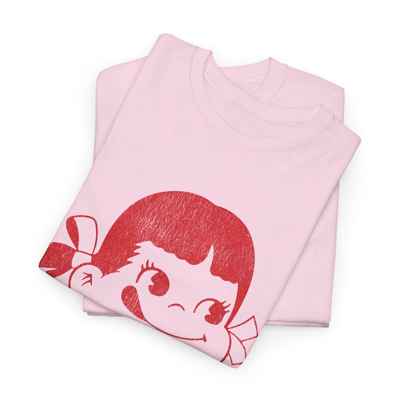 Two folded light pink t-shirts, with the top shirt displaying a red graphic of a winking cartoon girl's face with a bow in her hair.