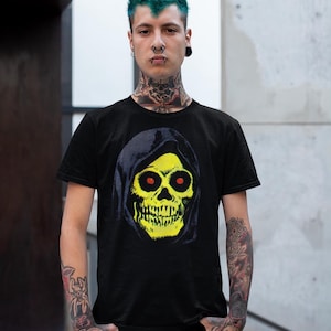 A person with green hair and tattoos wears a black t-shirt with a yellow skull in a blue hood, standing in front of a concrete wall.