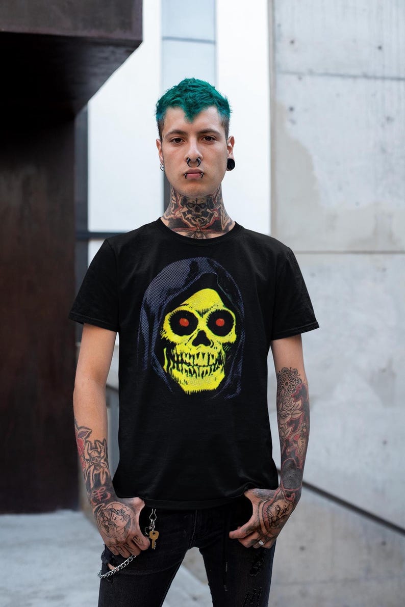 A person with green hair and tattoos wears a black t-shirt with a yellow skull in a blue hood, standing in front of a concrete wall.