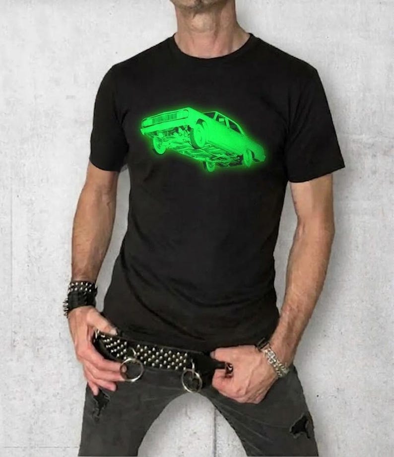 May include: A black short-sleeved t-shirt featuring a bright neon green graphic of a classic car on the chest. The shirt is paired with dark gray distressed jeans and a wide black belt adorned with silver metal studs and two large metal rings.