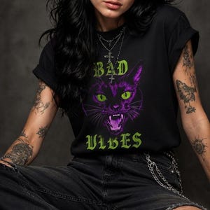 May include: A black short-sleeved t-shirt featuring a graphic of a purple cat with bright green eyes and an open mouth. The text "BAD" appears above the cat and "VIBES" appears below in a green gothic font. The shirt has a crew neckline.