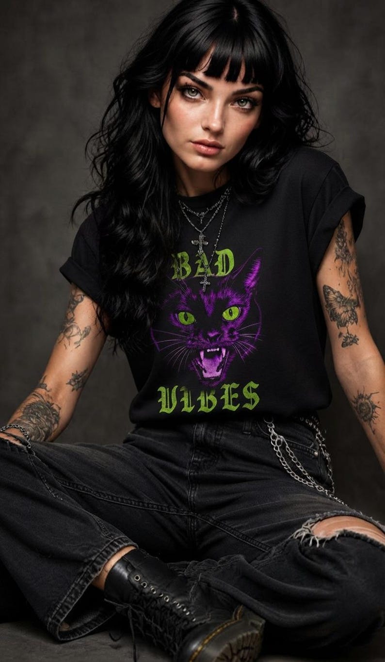 May include: A black short-sleeved t-shirt featuring a graphic of a purple cat with bright green eyes and an open mouth. The text "BAD" appears above the cat and "VIBES" appears below in a green gothic font. The shirt has a crew neckline.
