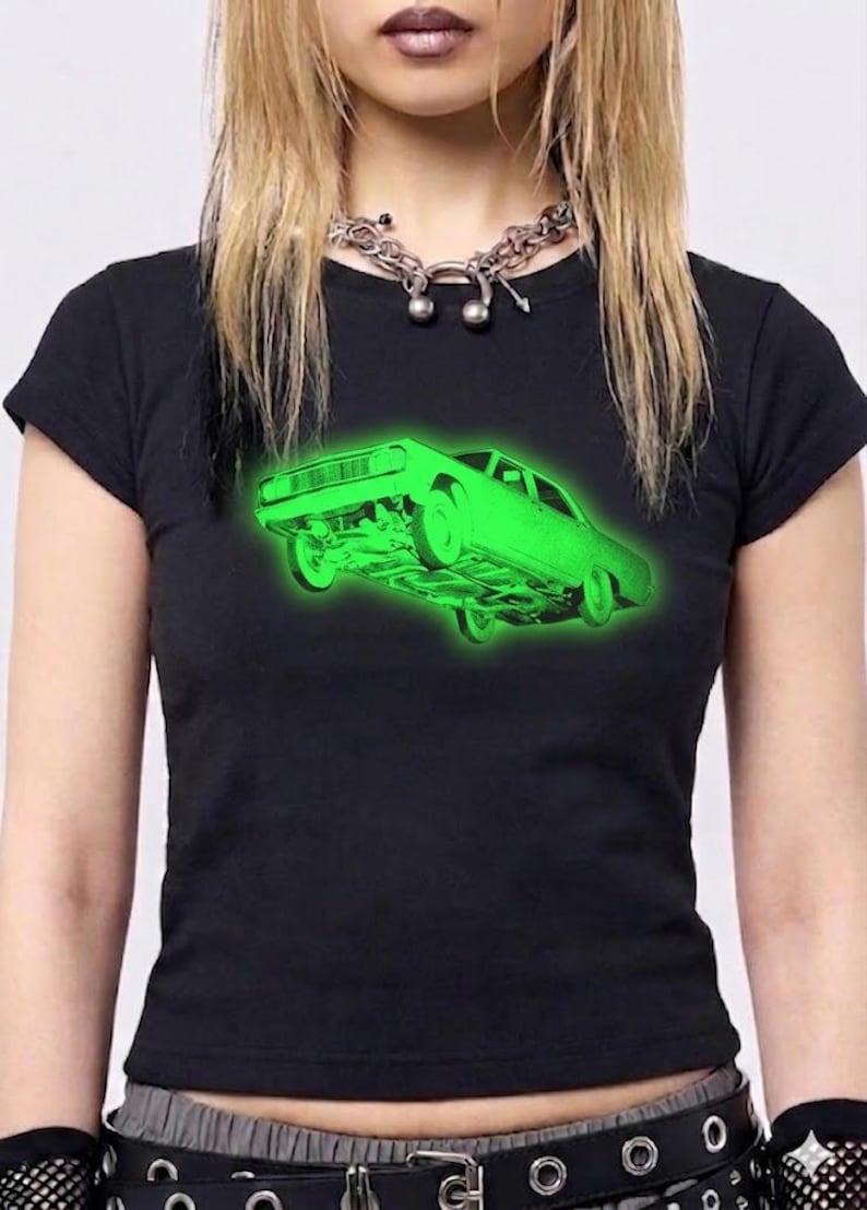 May include: A black short-sleeved t-shirt featuring a vibrant neon green graphic of a car floating in the center. The car illustration shows the underside and wheels in detail, creating a glowing effect against the dark fabric of the shirt.