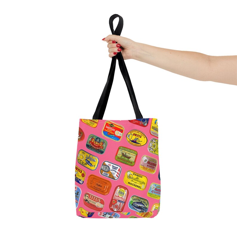 A hand holds up a pink tote bag patterned with colorful sardine can labels, against a solid yellow background.