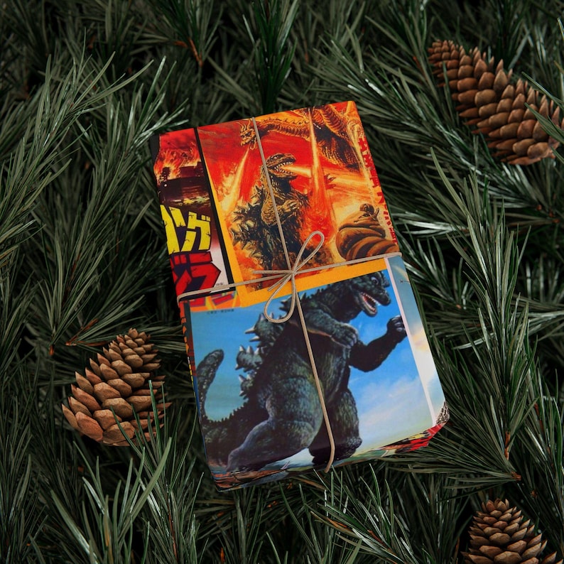 A small rectangular gift wrapped in Godzilla-themed paper, tied with twine, resting on green pine needles with pinecones.