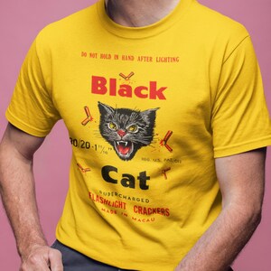 May include: Yellow t-shirt featuring a black cat graphic with red accents and the text "Black Cat Flashlight Crackers". The shirt also has the text "Do not hold in hand after lighting".