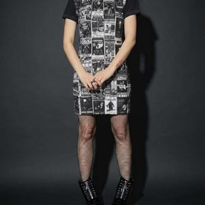 A full-length view of a model standing in a black t-shirt dress with a vintage horror poster print, fishnet tights, and black lace-up boots on a dark background.