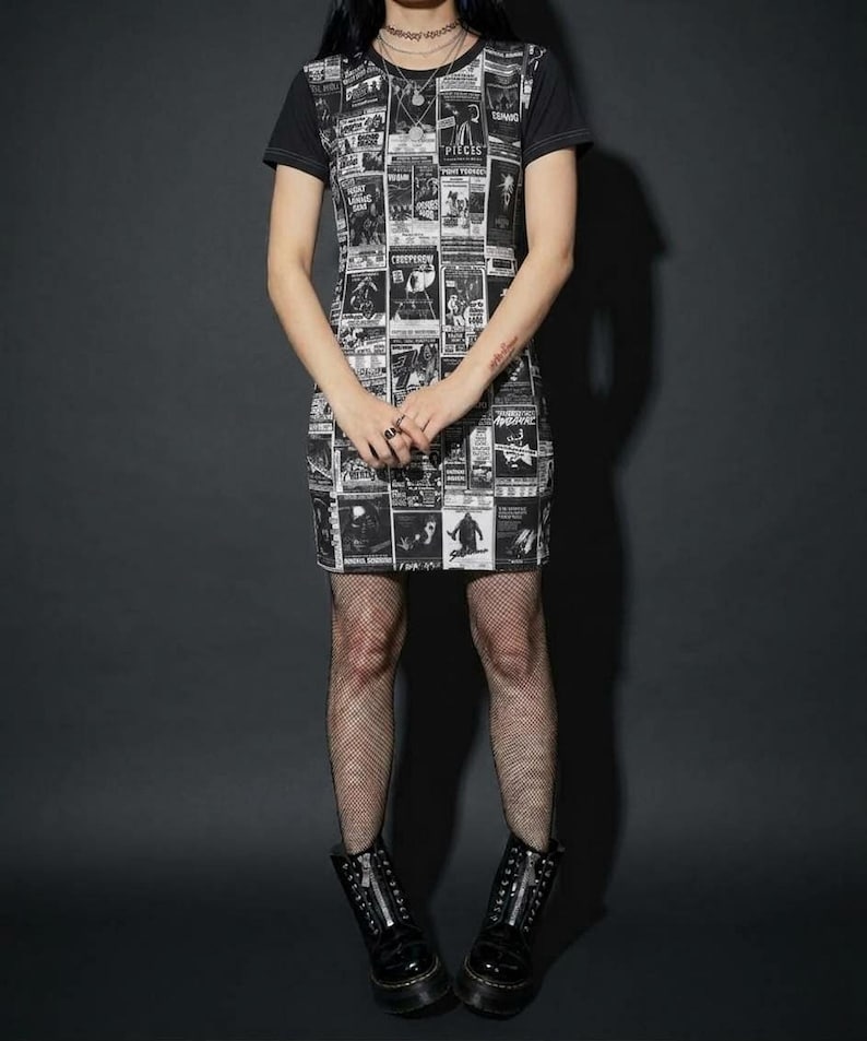A full-length view of a model standing in a black t-shirt dress with a vintage horror poster print, fishnet tights, and black lace-up boots on a dark background.