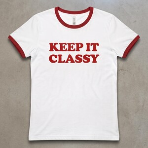 May include: A white ringer t-shirt with short sleeves and a red crew neckline. The shirt features the text KEEP IT CLASSY printed in bold red capital letters across the center of the chest. The sleeve cuffs are also trimmed in red.