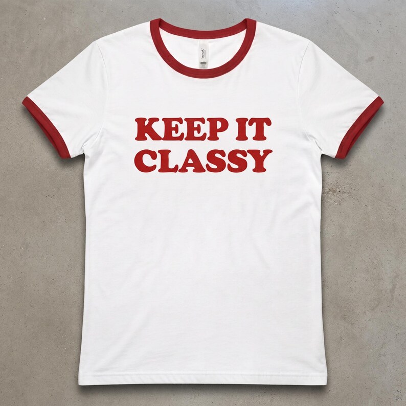 May include: A white ringer t-shirt with short sleeves and a red crew neckline. The shirt features the text KEEP IT CLASSY printed in bold red capital letters across the center of the chest. The sleeve cuffs are also trimmed in red.