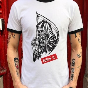 A person wearing a white ringer t-shirt with black collar and sleeve trim, featuring a black illustration of a grim reaper holding a scythe above a red banner with the text Killin it.