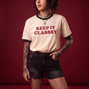 May include: A cream-colored ringer t-shirt with black trim on the collar and sleeves, featuring the text "KEEP IT CLASSSY" in red lettering, paired with black denim shorts that have a distressed, frayed hem and a button-fly closure.