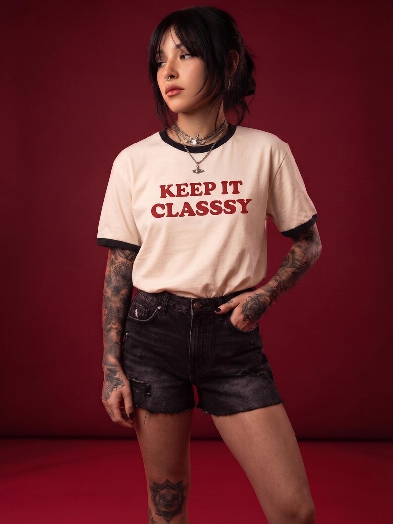 May include: A cream-colored ringer t-shirt with black trim on the collar and sleeves, featuring the text "KEEP IT CLASSSY" in red lettering, paired with black denim shorts that have a distressed, frayed hem and a button-fly closure.