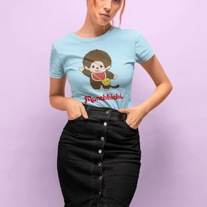 May include: A light blue short-sleeved t-shirt featuring a graphic of a brown Monchhichi character wearing a red bib and a yellow bag, with the red text Monchhichi printed below. The shirt is paired with a black denim skirt with a front button closure.