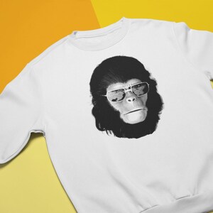 K&ouml;nnte beinhalten: Wei&szlig;es Sweatshirt mit einer schwarz-wei&szlig;en Grafik eines Affen mit Sonnenbrille. Das Gesicht des Affen ist auf der Vorderseite des Rundhals-Oberteils abgebildet. Der Hintergrund ist gelb und orange.