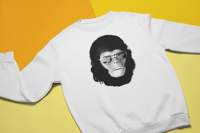K&ouml;nnte beinhalten: Wei&szlig;es Sweatshirt mit einer schwarz-wei&szlig;en Grafik eines Affen mit Sonnenbrille. Das Gesicht des Affen ist auf der Vorderseite des Rundhals-Oberteils abgebildet. Der Hintergrund ist gelb und orange.