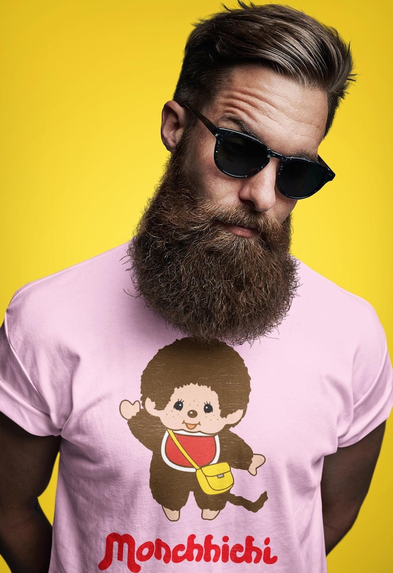 A man with a large beard and sunglasses wears a light pink t-shirt featuring a Monchhichi graphic against a yellow background.
