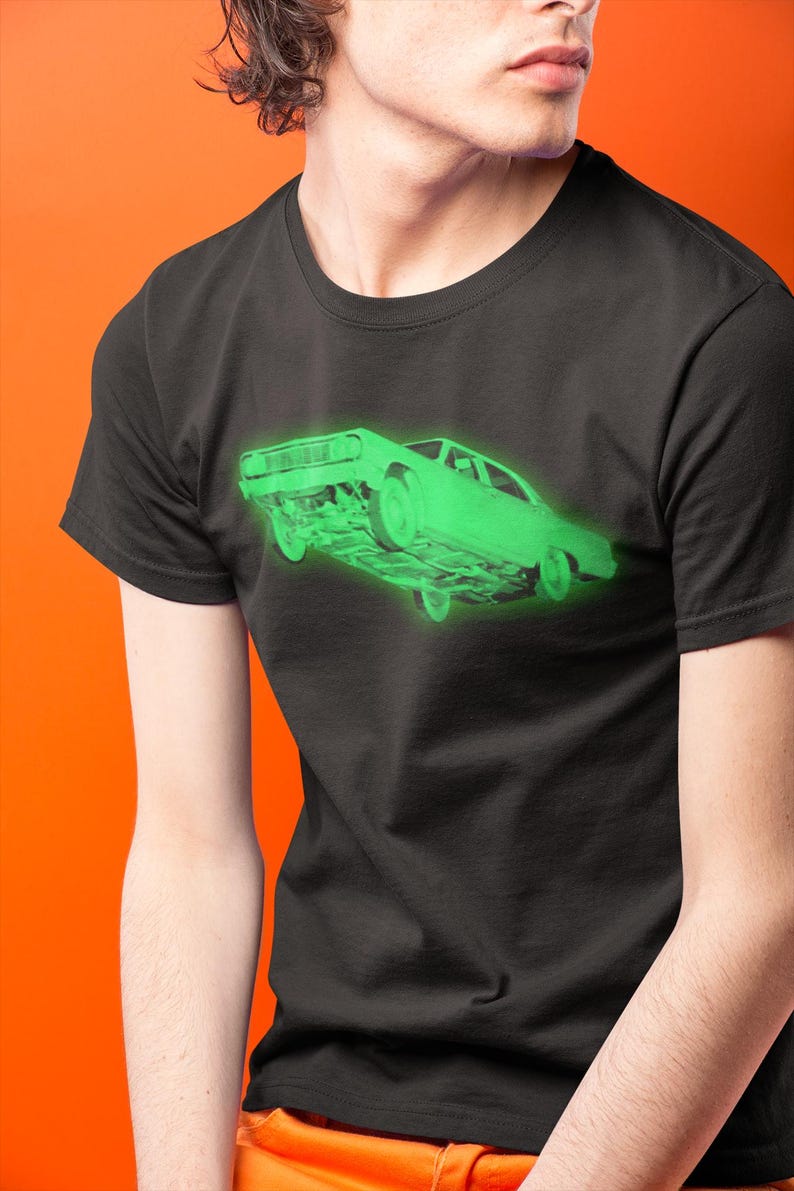May include: A black short-sleeved t-shirt featuring a glowing neon green graphic of a vintage car positioned at an angle. The car illustration shows the front and side profile with a bright, luminous effect against the dark fabric of the shirt.