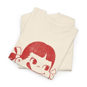Two folded natural-colored t-shirts, with the top shirt displaying a red graphic of a winking cartoon girl's face with a bow in her hair.