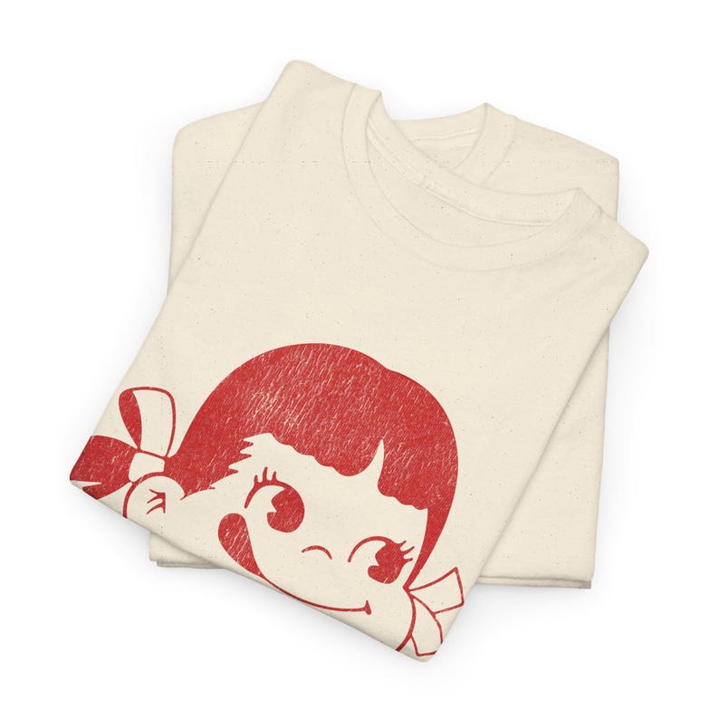Two folded natural-colored t-shirts, with the top shirt displaying a red graphic of a winking cartoon girl's face with a bow in her hair.