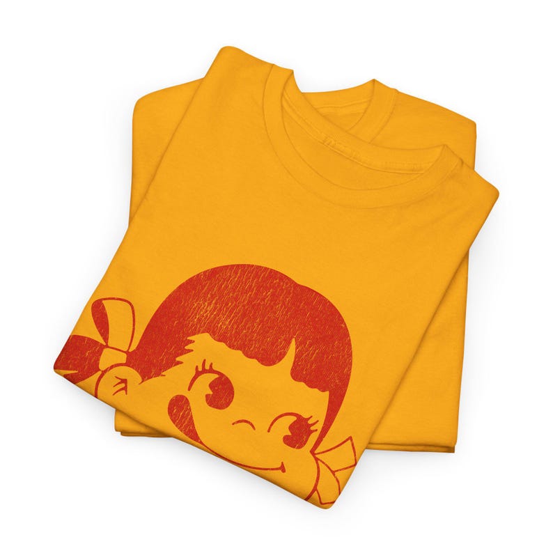 Two folded mustard yellow t-shirts, with the top shirt displaying a red graphic of a winking cartoon girl's face with a bow in her hair.