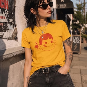 A person with dark hair and sunglasses wears a mustard yellow t-shirt with a red graphic of a girl's face with pigtails and a winking expression, paired with black denim shorts, leaning against a wall outdoors.