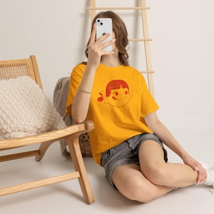 A person sits on a wooden floor, holding a phone to their face, wearing a mustard yellow t-shirt with a red graphic of a girl's face with pigtails and a winking expression, paired with dark shorts.