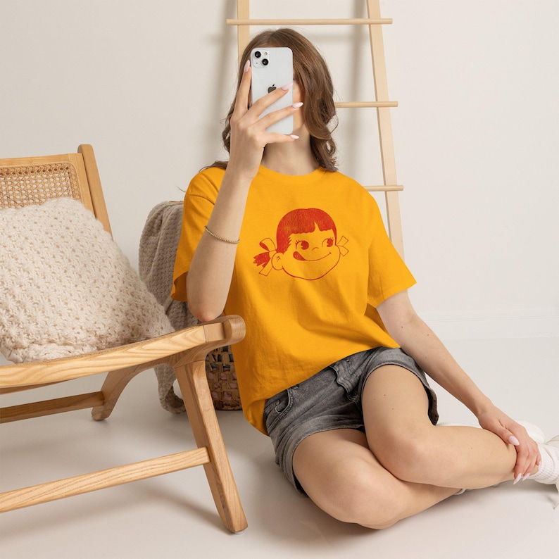 A person sits on a wooden floor, holding a phone to their face, wearing a mustard yellow t-shirt with a red graphic of a girl's face with pigtails and a winking expression, paired with dark shorts.