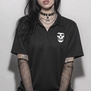 A person with dark hair wearing a black polo shirt featuring a white skull embroidery on the chest, layered necklaces, and fishnet sleeves against a neutral background.
