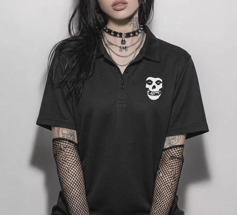 A person with dark hair wearing a black polo shirt featuring a white skull embroidery on the chest, layered necklaces, and fishnet sleeves against a neutral background.