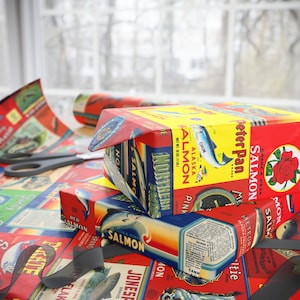 May include: A stack of vintage-style salmon can labels wrapped around boxes. The labels feature red, yellow, and blue colors with illustrations of salmon and text that reads "Grade A KETA SALMON", "Peter Pan Brand", "Alaska Salmon", "Northern Pride", "Pink Salmon", "Meteor Salmon", "Jones Bros. Brand", "Red Salmon", "Fishnelak", "Kate", "DanTy", and "Mon".